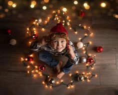 a young boy is sitting on the floor surrounded by christmas ornaments and lights, looking up at the camera