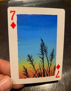 a hand holding up a playing card with the image of grass in front of an orange and blue sky