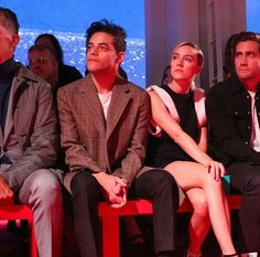 three people sitting on red benches in front of an audience at a fashion show, with one person looking up