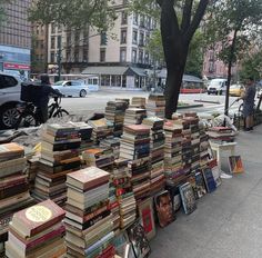 many books are stacked up on the side of the road near a tree and sidewalk