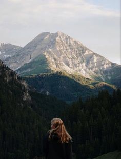 a woman standing on top of a lush green hillside next to a forest covered mountain