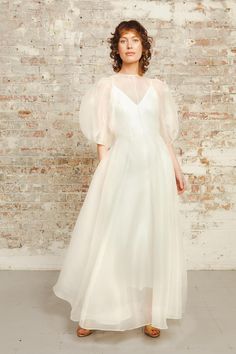 a woman standing in front of a brick wall wearing a white dress with sheer sleeves