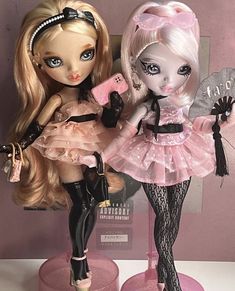 two dolls that are standing next to each other on a table with pink walls behind them