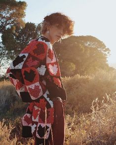 a woman standing in a field wearing a red and black crocheted jacket with hearts on it