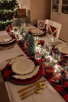 the table is set for christmas dinner with red and white plates, silverware, and evergreen trees