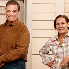 an older man and woman standing in front of a house with their hands on their hips