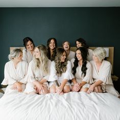 a group of women sitting on top of a bed in white robes smiling at the camera