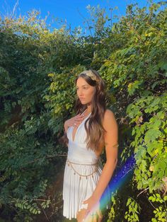 a woman in a white dress standing next to some bushes and trees with her hands on her hips