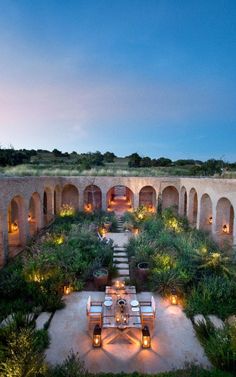 an outdoor dining area is lit up with candles and greenery at dusk, surrounded by stone arches