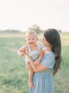 a woman holding a baby in her arms and smiling at each other while standing on the grass