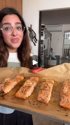 a woman is holding a tray with salmon on it and looking at the camera while wearing glasses