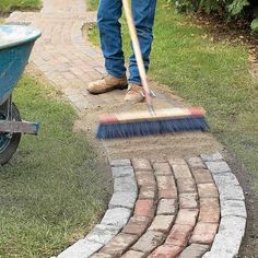 a man with a broom is cleaning the grass near a brick walkway and wheelbarrow