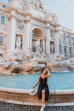 a woman sitting on the edge of a fountain in front of a building with statues