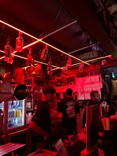 people sitting at tables in a bar with red lights on the ceiling and bottles hanging from the ceiling