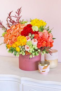 an arrangement of colorful flowers in a pink vase on a white dresser against a pink wall