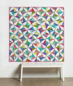 a colorful quilt is hanging on the wall next to a white stand with a small stool