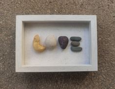 three rocks in a white shadow box on the ground, one with a yellow banana and one with a red heart