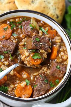 a white bowl filled with beef and barley stew next to rolls on the side,