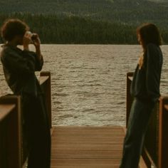 two people standing on a dock near the water taking pictures with their cell phones,