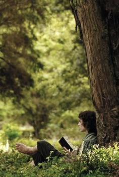 a man sitting under a tree reading a book in the woods with his legs crossed