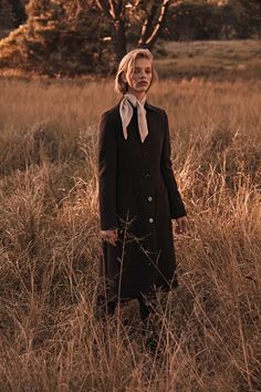 a woman standing in the middle of a field wearing a black coat and white shirt