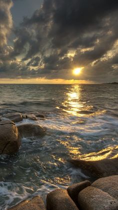 the sun is setting over the ocean with rocks in the foreground and dark clouds above