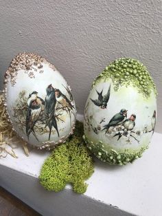 three decorated eggs sitting on top of a white shelf next to green moss covered plants