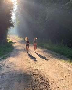 two people running down a dirt road in the sun with trees and bushes behind them