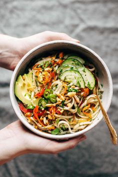 a person holding a bowl of food with noodles, cucumbers and other vegetables