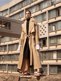 a woman standing in front of a building wearing a tan trench coat and black dress