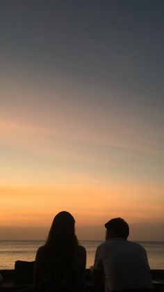 two people sitting on a bench watching the sun set over the ocean in front of them