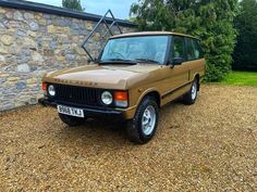 a brown jeep parked in front of a stone wall and grass covered driveway with trees behind it