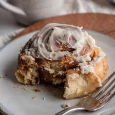 a cinnamon roll with icing on a white plate next to a fork and cup