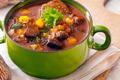 a green bowl filled with stew and vegetables on top of a white napkin next to bread