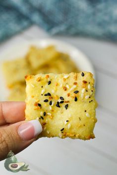 a hand holding up a piece of bread with sesame seeds on it and some other food in the background