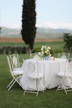 a table with white chairs and flowers on it in the middle of a grassy field