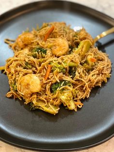 a black plate topped with noodles and veggies next to a silver serving spoon