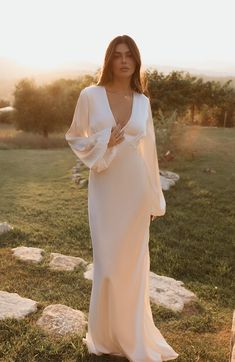 a woman standing in the grass wearing a long white dress with bell sleeves and an open back