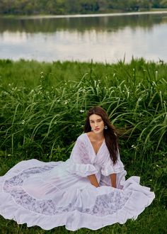 a woman is sitting on the grass in front of a lake wearing a white dress