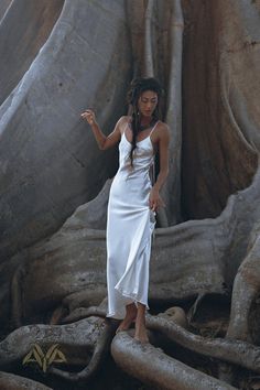 a woman in a white dress standing next to a large tree with its roots exposed