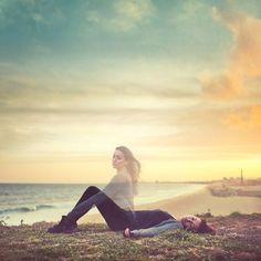 a man and woman sitting on the grass near the ocean at sunset or sunrise, looking into the distance