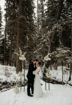 a bride and groom kissing in the snow surrounded by tall pine trees at their winter wedding