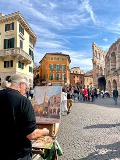 an older man is painting in front of some old buildings and people are walking around