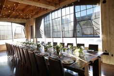 a long wooden table with lots of plates and vases on it in front of large windows