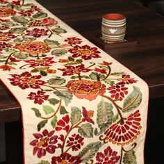 a table runner with flowers on it and a cup sitting next to it, in front of a wooden table