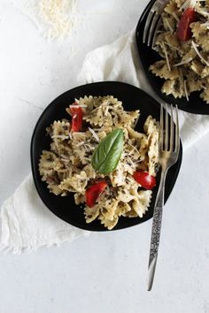 two black plates filled with pasta and vegetables on top of a white table cloth next to silverware