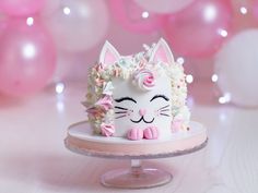 a cat cake on a plate with pink and white balloons in the backgroud