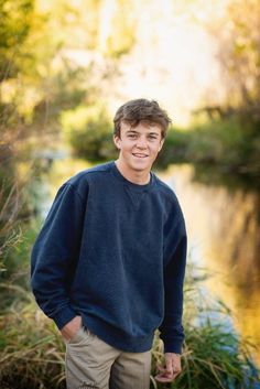 a young man standing in front of a body of water wearing a blue sweater and khaki pants