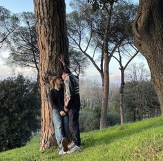 two people standing next to each other in the grass near some trees and one person is leaning against a tree