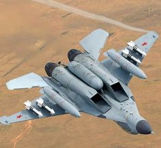 a fighter jet flying through the sky over a desert area with grass and trees in the background